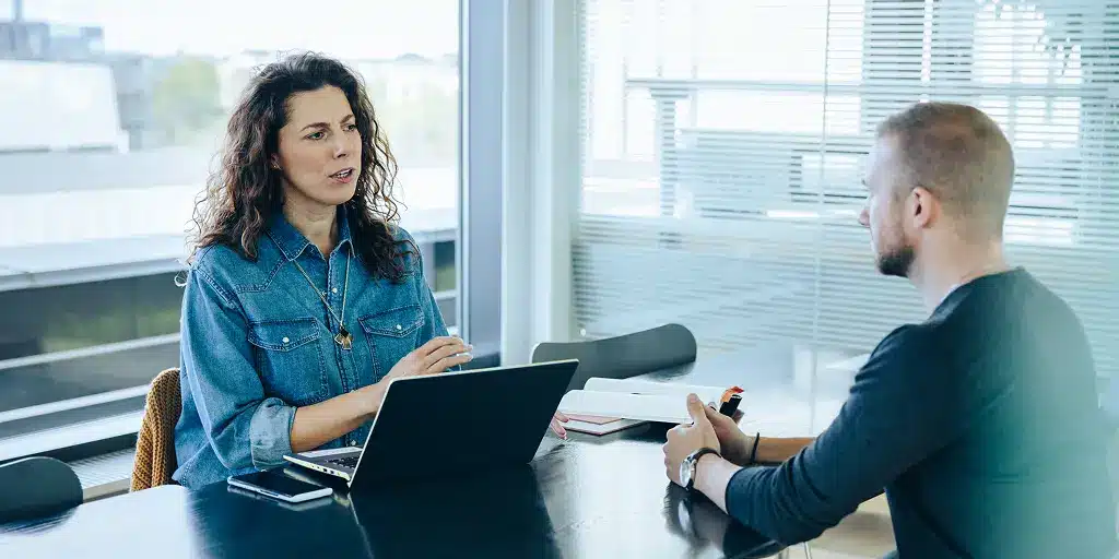 Interviewer:in führt ein Bewerbungsgespräch mit einem Bewerber im Büro und stellt typische Fragen im Bewerbungsgespräch.