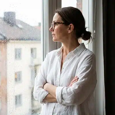 Eine Frau mit dunkel blonden Haaren, die Haare zu einem Zopf zusammen gebunden, schaut nachdenklich aus dem Fenster.