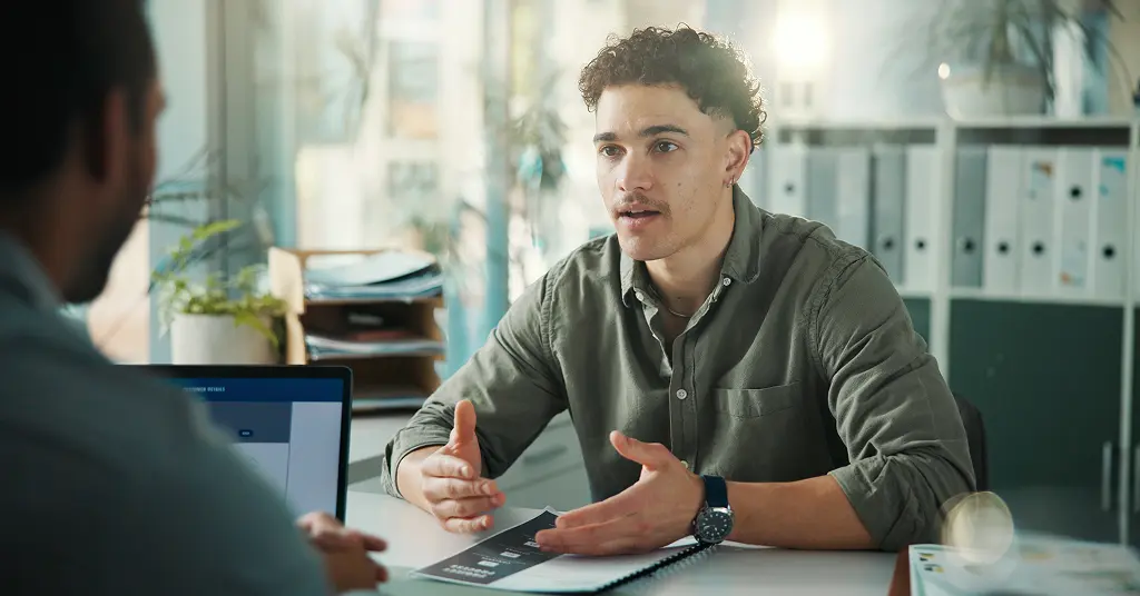 Junger Bewerber erklärt im Vorstellungsgespräch seine Stärken und Schwächen mit konkreten Beispielen aus der Praxis – gegenüber sitzt ein Interviewer mit Laptop
