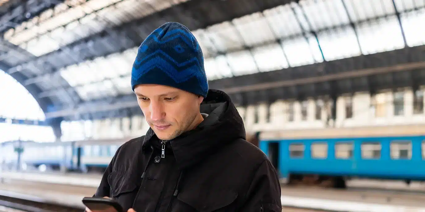 Mann mit Mütze und Winterjacke steht an einem Bahnhof und schaut auf sein Smartphone – AVGS Coaching Berlin für internationale Fachkräfte, die in Deutschland beruflich durchstarten möchten.